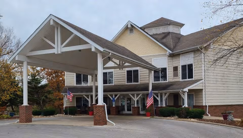 Exterior view of St. Andrew's Apartments at Kirksville showing a two-story building with beige siding and a covered entrance supported by white pillars with brick bases. There are American flags on either side of the entrance and some trees with autumn foliage in the background.