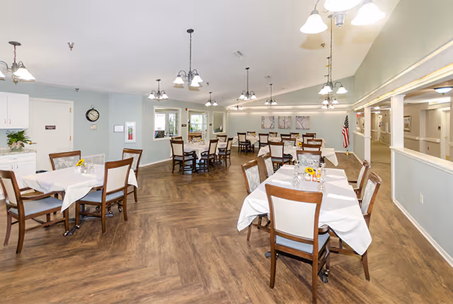 A spacious dining room with several tables covered in white tablecloths and wooden chairs arranged around them. The room has wood-patterned flooring, soft green walls, and multiple ceiling light fixtures. There is an American flag in the corner and a hallway visible to the right.