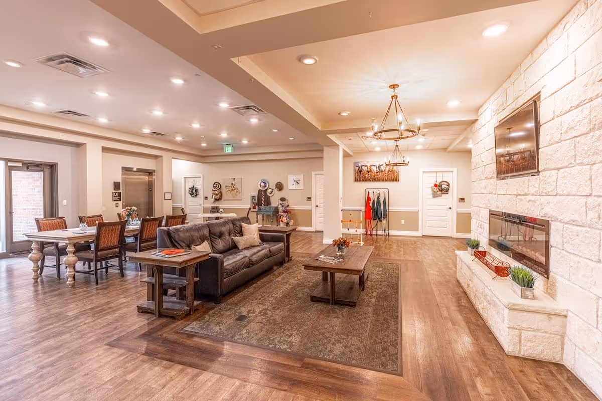 A spacious and well-lit common area in an assisted living facility featuring a brown leather sofa, a wooden coffee table on a patterned rug, a stone fireplace with a mounted TV above it, and a dining area with tables and chairs in the background. The room has wooden flooring, beige walls, and decorative lighting fixtures on the ceiling.