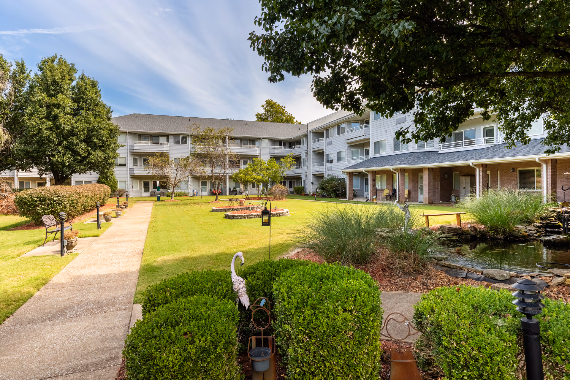 Outdoor courtyard area of Jackson Meadow - A Provincial Senior Living Community featuring a well-maintained lawn, trimmed bushes, a small pond with a water feature, benches along a paved walkway, and a multi-story residential building with balconies in the background under a partly cloudy sky.