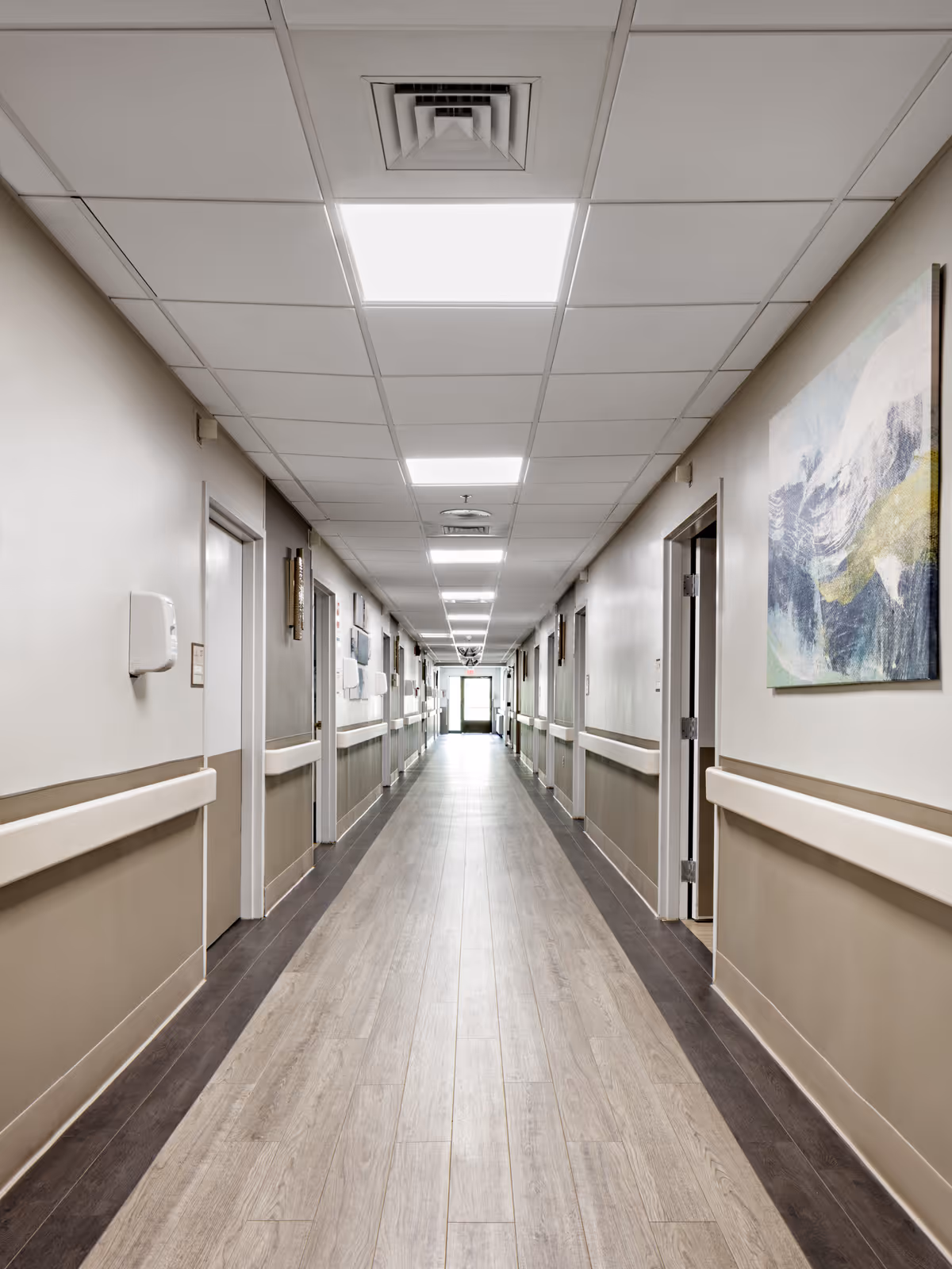 A long, well-lit hallway in a healthcare facility with light-colored walls, wood-patterned flooring, multiple closed doors on both sides, handrails along the walls, ceiling lights, and a piece of abstract artwork on the right wall.