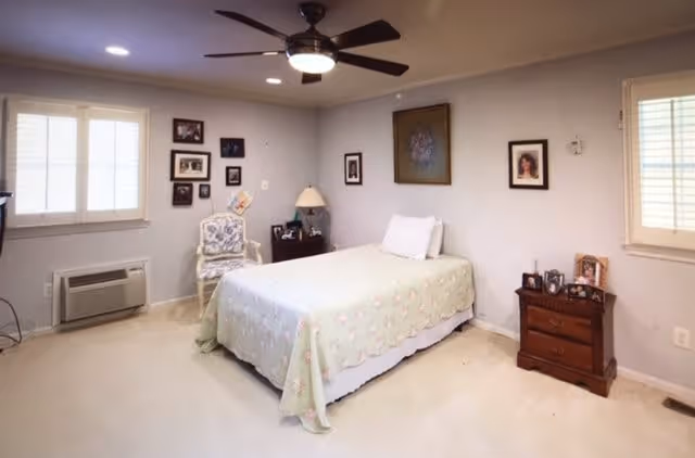 A bedroom with a single bed covered with a light green floral bedspread. The room has light-colored walls with several framed pictures hanging on them. There is a wooden nightstand with framed photos and a lamp on one side of the bed, and a white cushioned chair with a floral pattern near the corner. The room has two windows with white blinds and a ceiling fan with a light fixture.