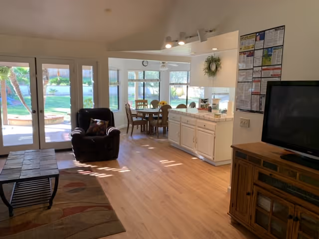 A bright living and dining area with wooden flooring. The living area has a dark armchair and a coffee table on a patterned rug. The dining area features a round wooden table with chairs and large windows letting in natural light. A kitchen counter with white cabinets separates the dining area from the living space. A TV sits on a wooden cabinet to the right, and glass doors open to a green outdoor space.