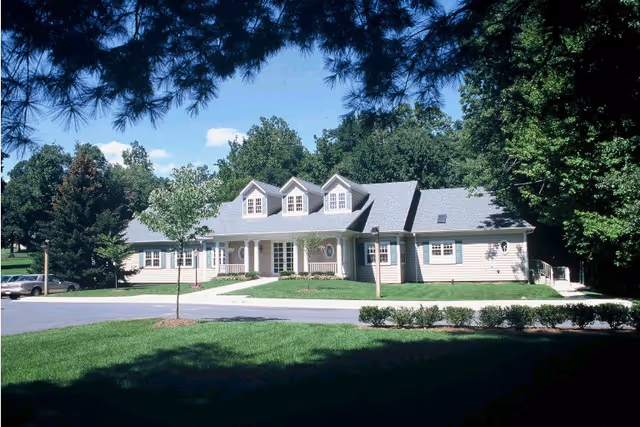 A single-story building with a gray roof and beige siding surrounded by green trees and a well-maintained lawn under a clear blue sky.