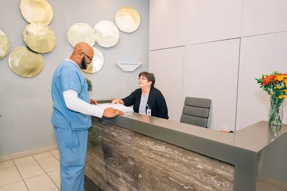 A man in blue medical scrubs stands at a reception desk speaking with a seated woman wearing glasses and a black cardigan. The reception area features a modern design with a stone front desk, a vase of colorful flowers on the counter, and decorative gold circular wall art in the background.