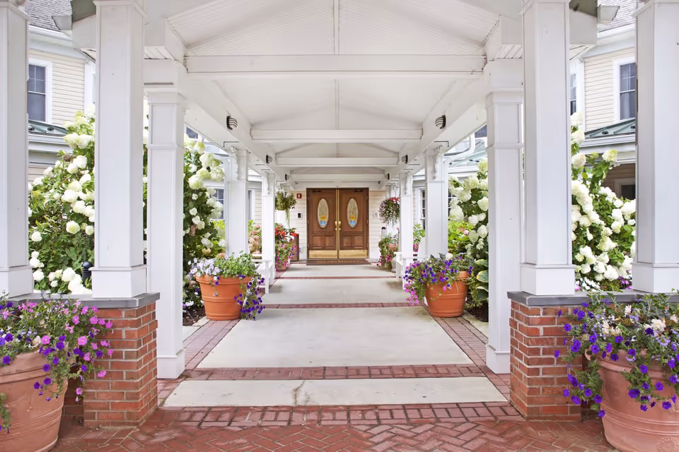 Covered white-columned entrance walkway lined with potted flowers and hydrangea bushes leading to double wooden doors.