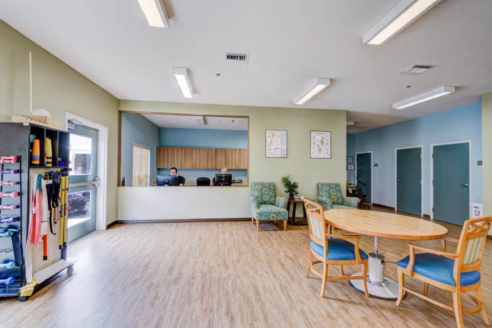 A bright and spacious room with light wood flooring and pastel green and blue walls. There is a reception window with two people working behind it, a round wooden table with four chairs, two patterned armchairs with a small table between them, and a rack with exercise equipment near a door with a window.