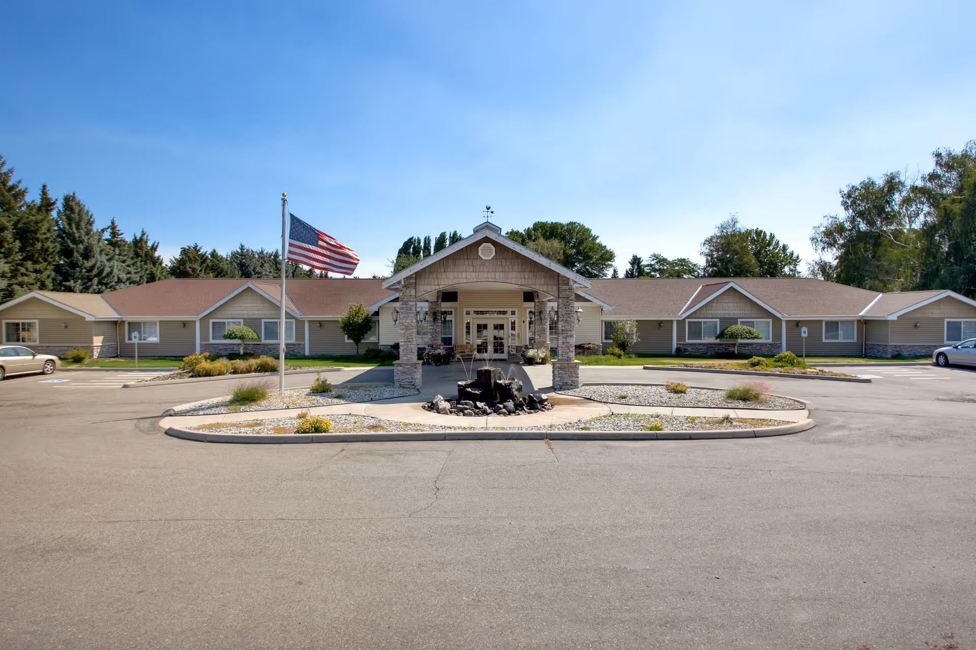 Front exterior view of Blossom Creek by Cogir facility with a circular driveway, an American flag on a flagpole, landscaped garden beds, and a covered entrance supported by stone pillars under a clear blue sky.