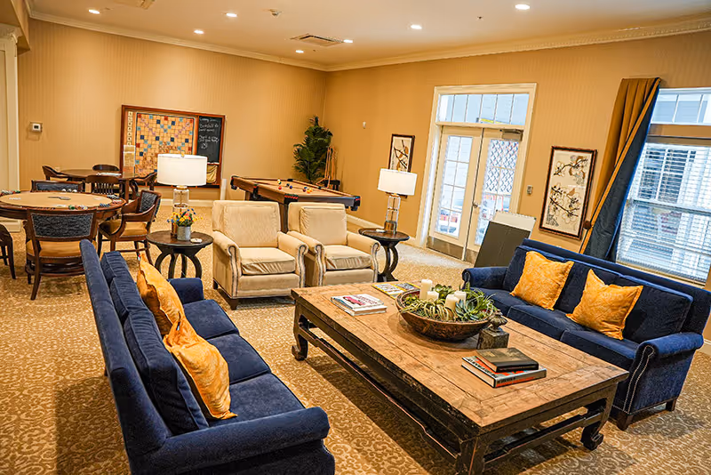 Spacious senior living common room with navy sofas, armchairs, a large wooden coffee table, game and card tables, and a pool table.