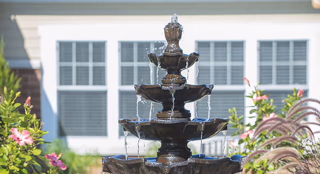 A multi-tiered black water fountain with water flowing from the top tier down to the lower tiers, surrounded by flowering plants and greenery, with a building featuring white walls and multiple windows in the background.