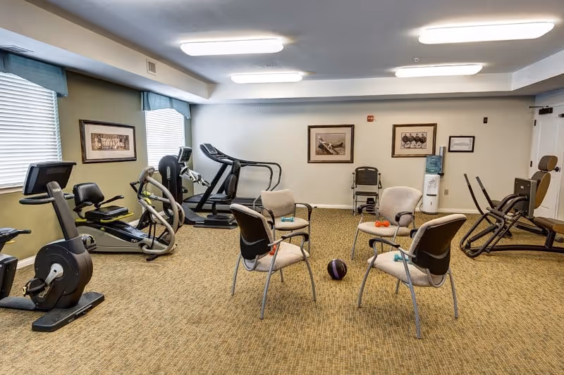 A senior living facility exercise room with various fitness equipment including stationary bikes, a treadmill, and weight machines. Four chairs are arranged in a circle in the center of the room with small exercise weights and a medicine ball on the floor. The room has carpeted flooring, framed pictures on the walls, and two large windows with blinds.