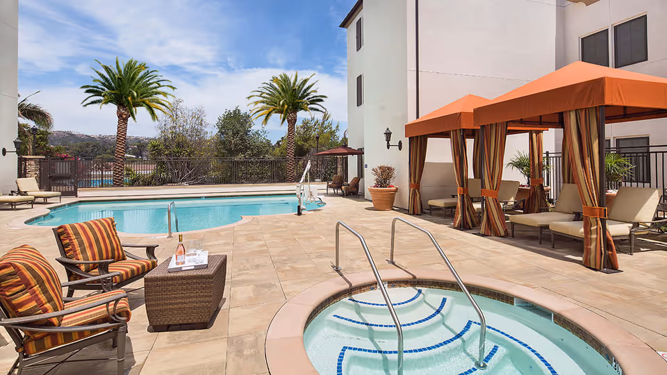 Outdoor pool area at Crestavilla featuring a swimming pool, a hot tub with handrails, several lounge chairs with striped cushions, a wicker table with a bottle and glasses, and cabanas with orange canopies. Palm trees and a clear sky are visible in the background.