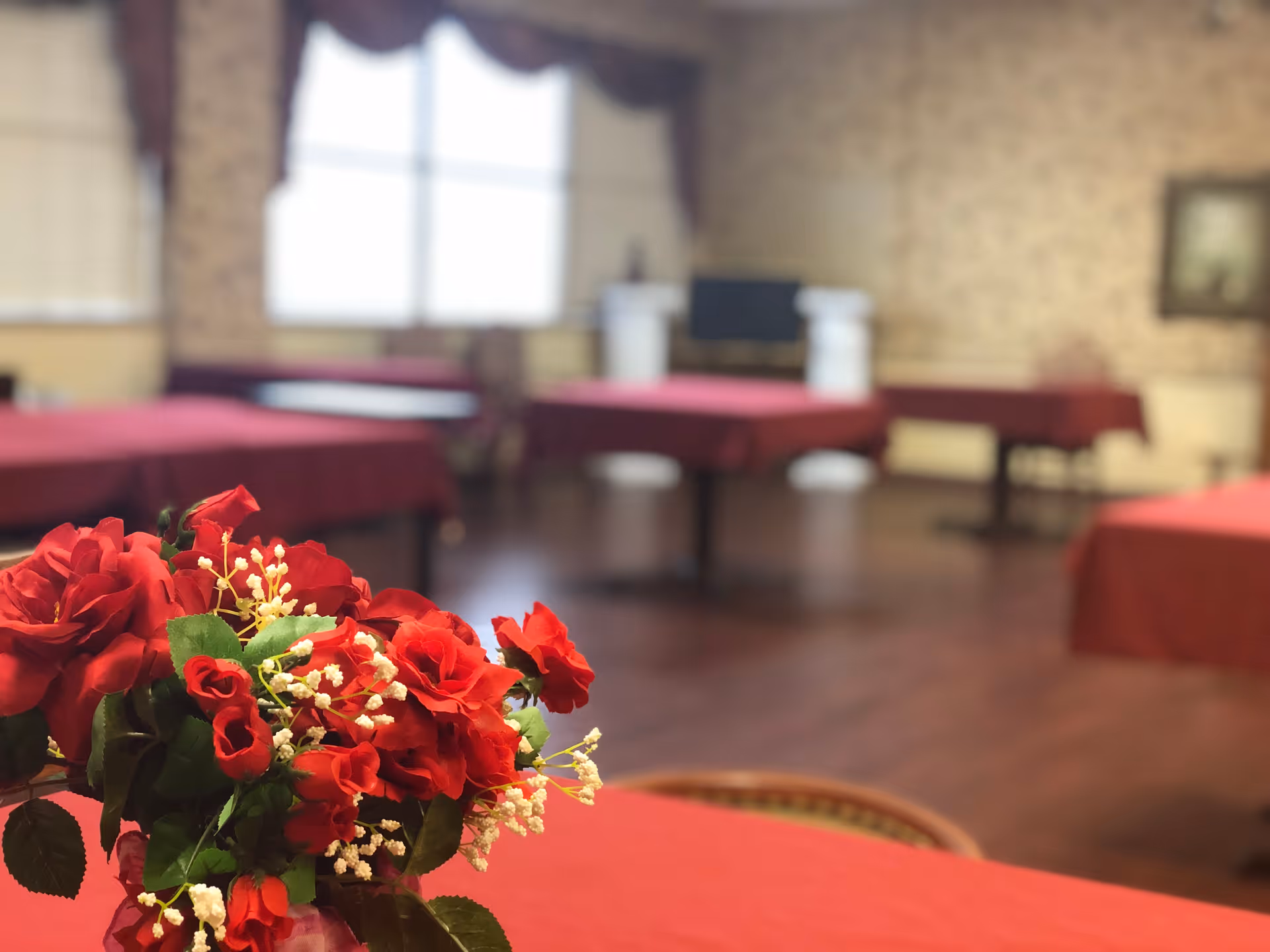 A dining room with tables covered in red tablecloths. In the foreground, there is a bouquet of red roses and white flowers. The room has wooden flooring, patterned wallpaper, and a window with curtains in the background.