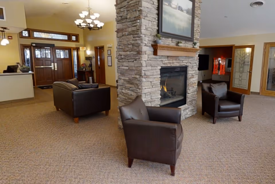 A cozy senior living facility common area with a stone fireplace in the center, two brown leather armchairs facing the fireplace, and a brown leather loveseat near the entrance. The room has beige carpet, warm yellow walls, wooden doors with glass panels, and a chandelier hanging from the ceiling.
