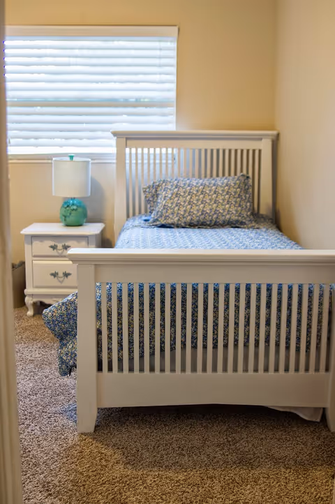 A small bedroom with a white wooden bed frame, a blue patterned bedspread and pillow, a white nightstand with two drawers, and a turquoise lamp. A window with white blinds is above the nightstand.