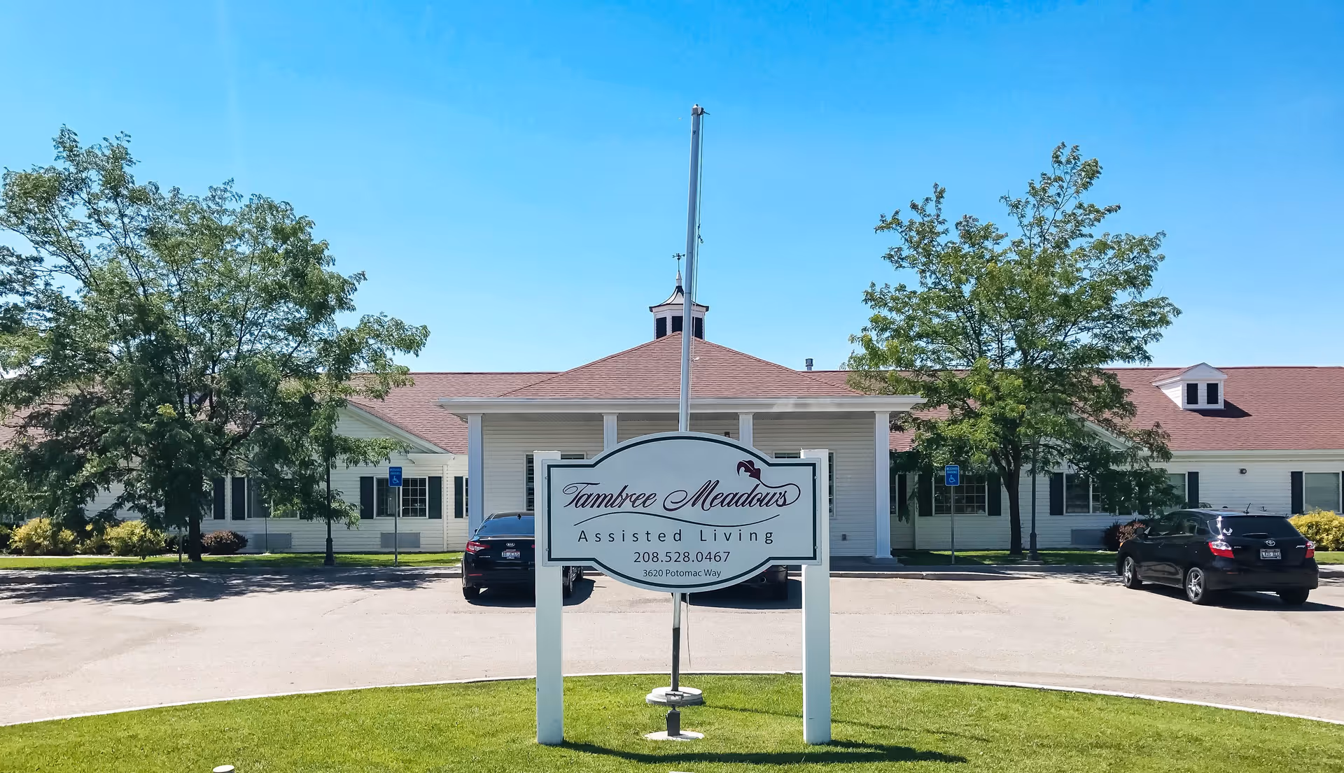 Exterior view of Tambree Meadows Assisted Living facility on a clear sunny day, showing a white building with a red roof, green trees, a parking area with cars, and a sign in the foreground displaying the facility name, phone number, and address.