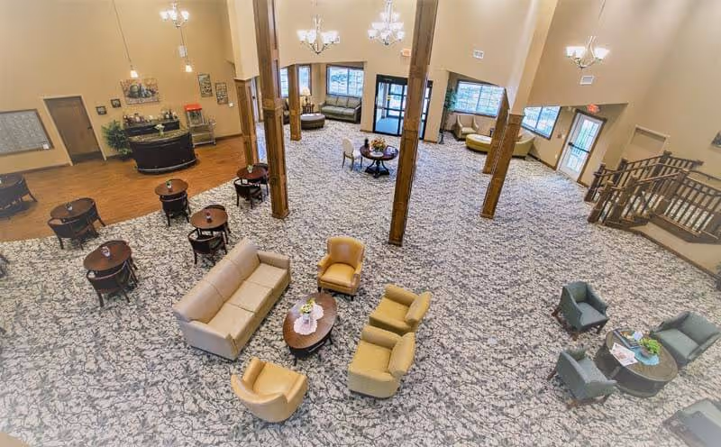 Spacious senior living facility common area with multiple seating arrangements including sofas, armchairs, and small tables. The room features carpeted flooring with a patterned design, wooden support beams, chandeliers, and large windows allowing natural light. There is a staircase on the right side and a reception or service counter area on the left.