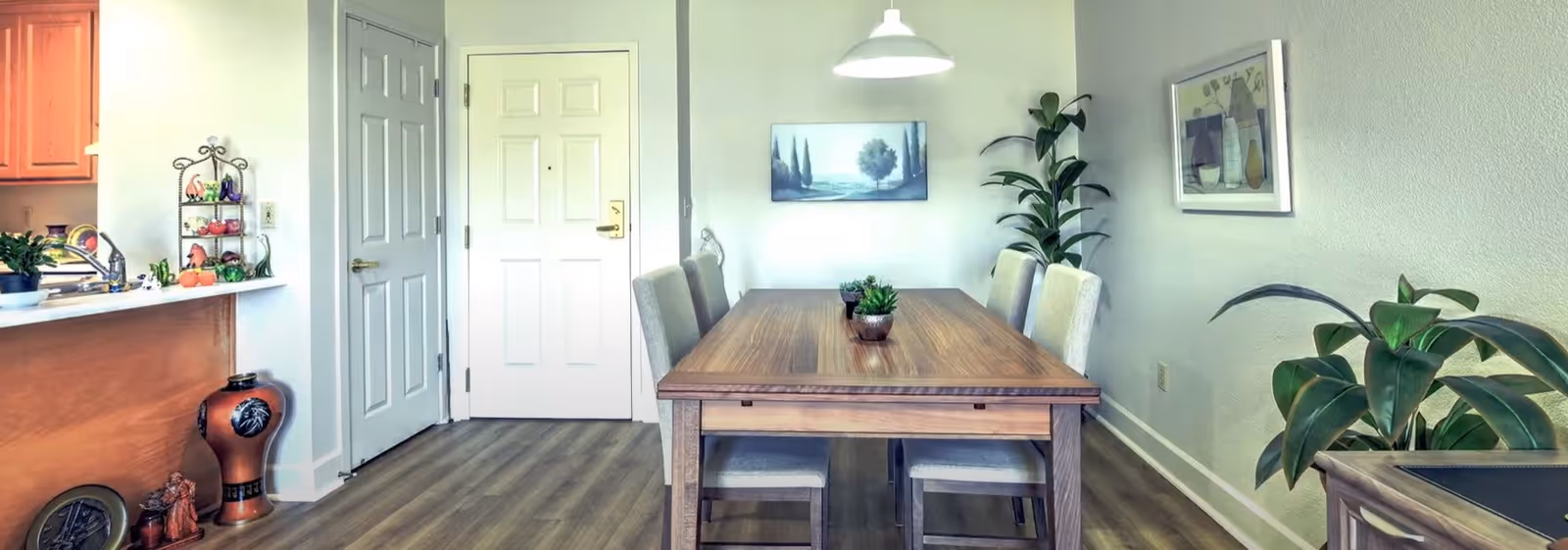 Bright dining room with a wooden table and four chairs, potted plants, wall art, and a view of the entry door and kitchen pass-through.