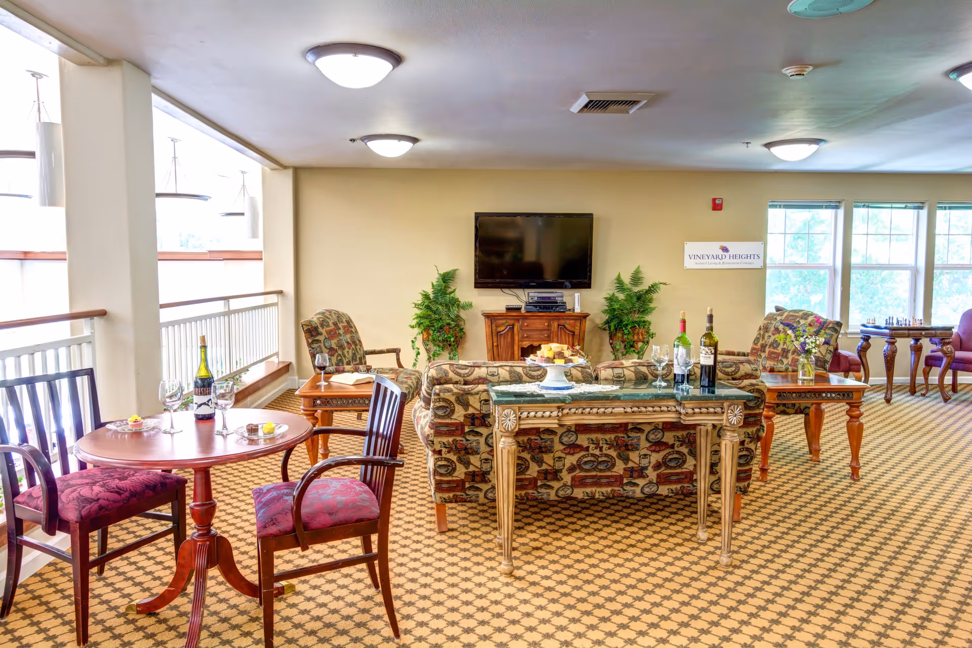 A bright assisted-living common room with patterned sofas and chairs, small tables set with wine glasses and bottles, a wall-mounted TV, and large windows.