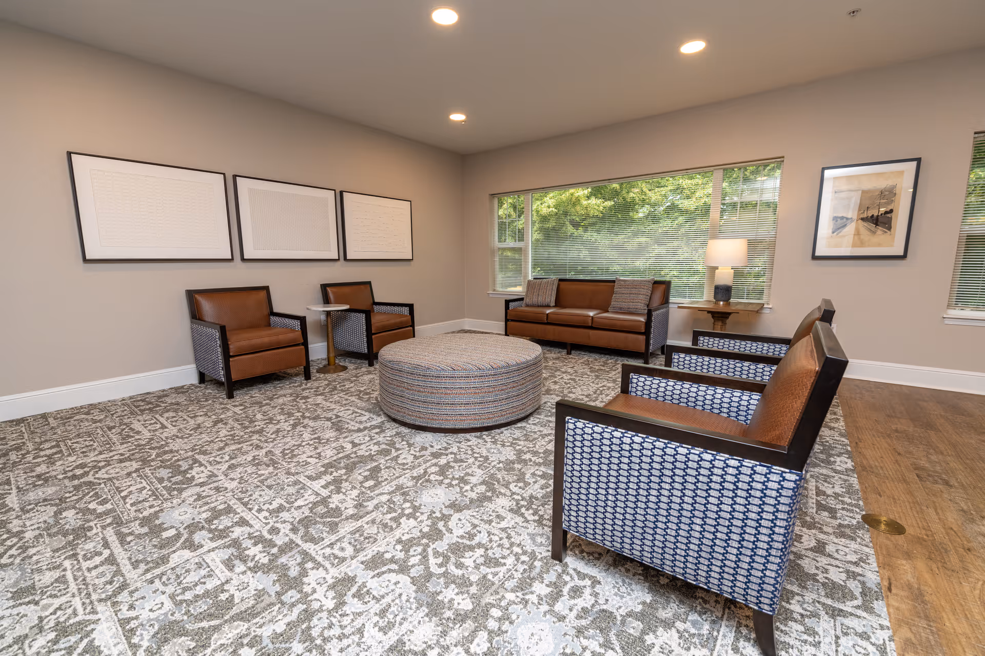 A spacious living room with a large patterned carpet, two brown leather armchairs with patterned fabric sides, a round upholstered ottoman in the center, a brown leather sofa with two pillows, a side table with a lamp, and large windows showing greenery outside. The walls are decorated with framed artwork.