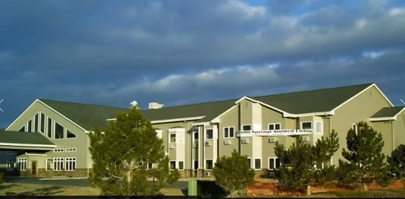 Exterior view of Autumn Springs Assisted Living facility, a two-story building with multiple windows, a green roof, and surrounded by trees under a cloudy sky.