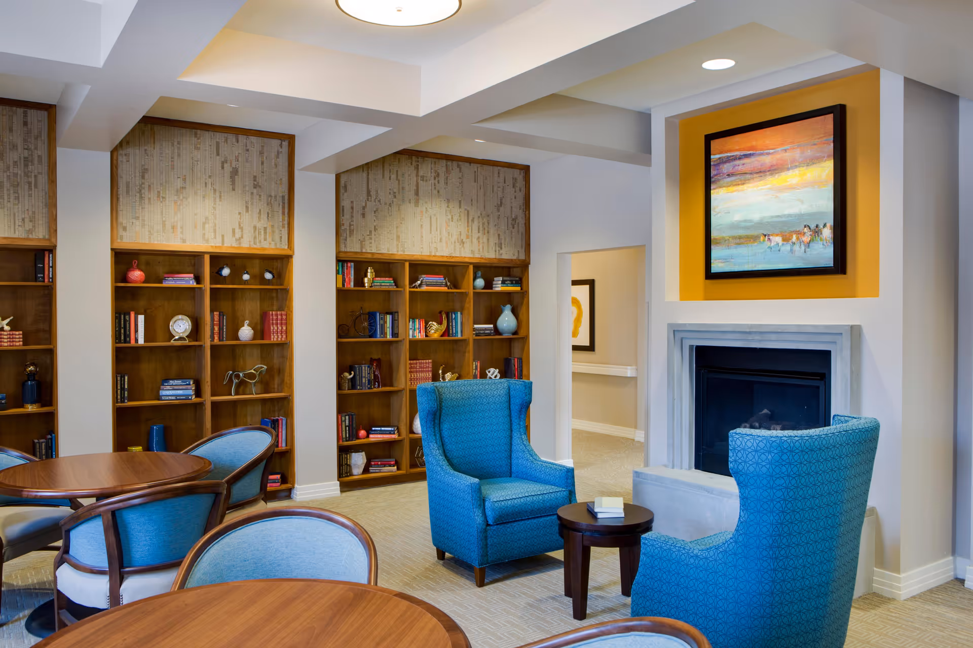 A cozy living room area in Savanna House featuring two blue upholstered armchairs facing a small round wooden table and a fireplace with a colorful abstract painting above it. Behind the chairs, there are wooden bookshelves filled with books and decorative items. The room has a warm and inviting atmosphere with soft lighting and neutral-colored walls.
