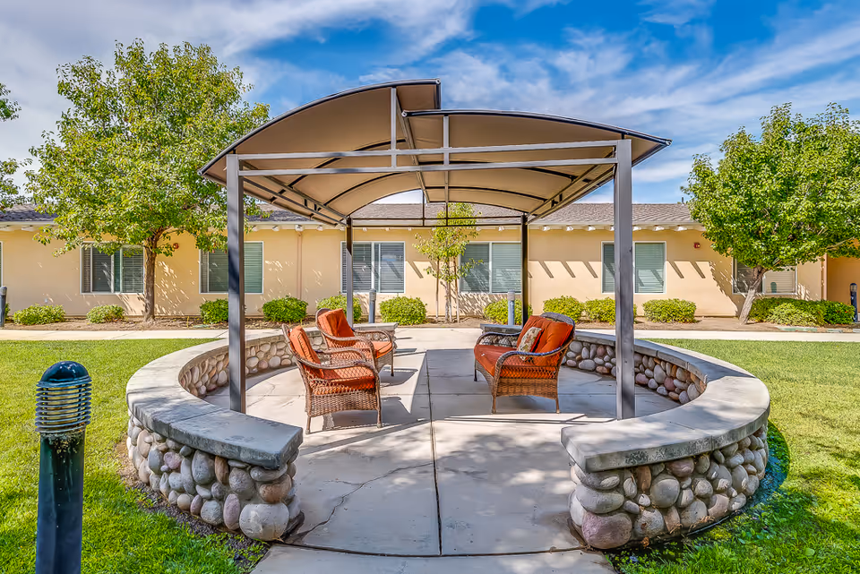 Outdoor seating area at Sunny Rose Senior Living featuring a circular stone wall with a metal canopy overhead. Inside the circle are cushioned wicker chairs and a loveseat arranged on a concrete patio. The background shows a single-story building with windows and green trees under a partly cloudy blue sky.