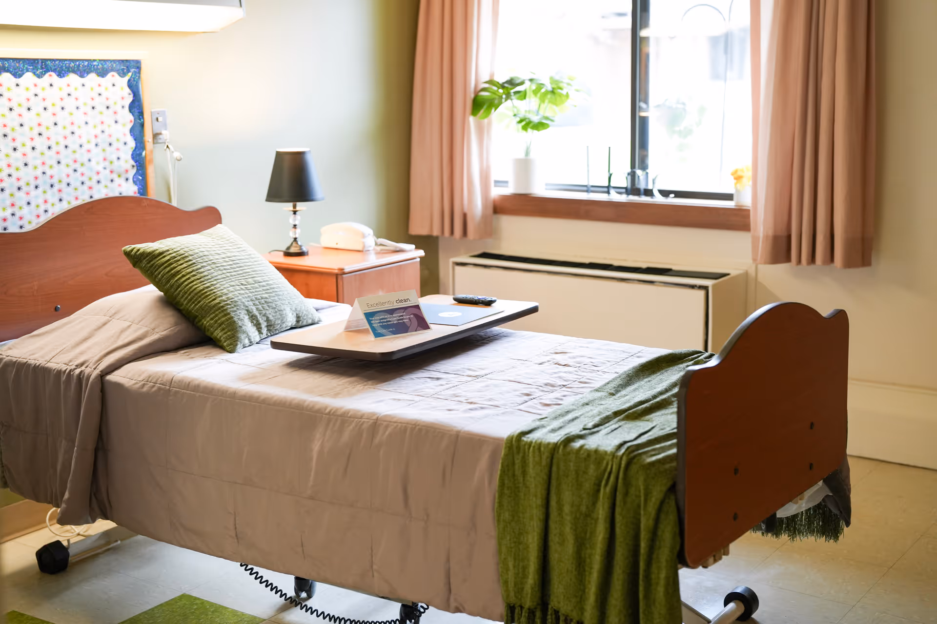 A neatly made hospital-style bed with a green pillow and green blanket in a well-lit room. A bedside table with a black lamp and a white telephone is next to the bed. A window with beige curtains and a potted plant on the windowsill is visible in the background. A tray with a card and a remote control rests on the bed.