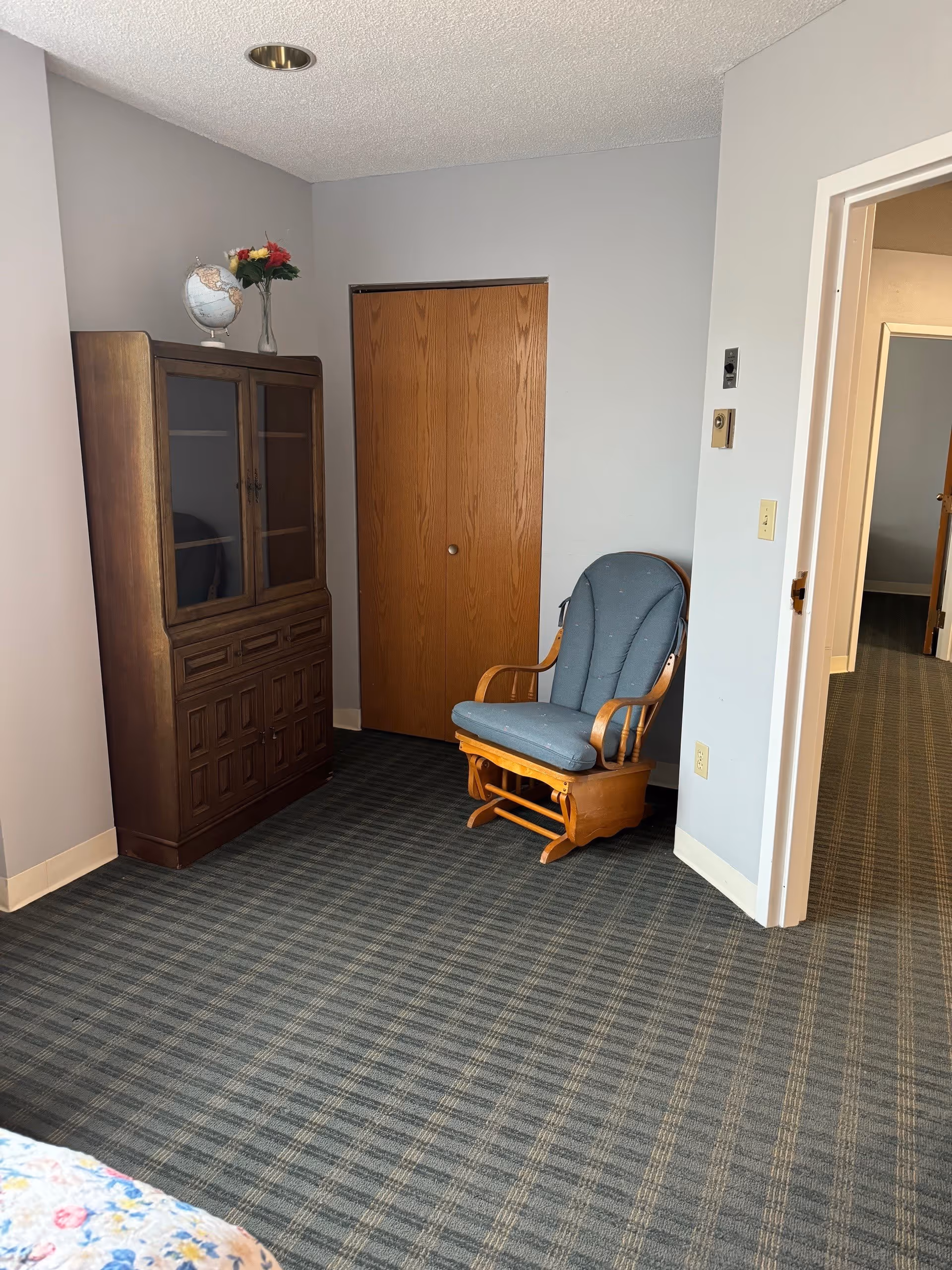 A corner of a room with a wooden rocking chair with blue cushions, a wooden cabinet with glass doors and drawers, a globe and a vase with flowers on top, a closed wooden closet door, and a doorway leading to another room with similar carpet.