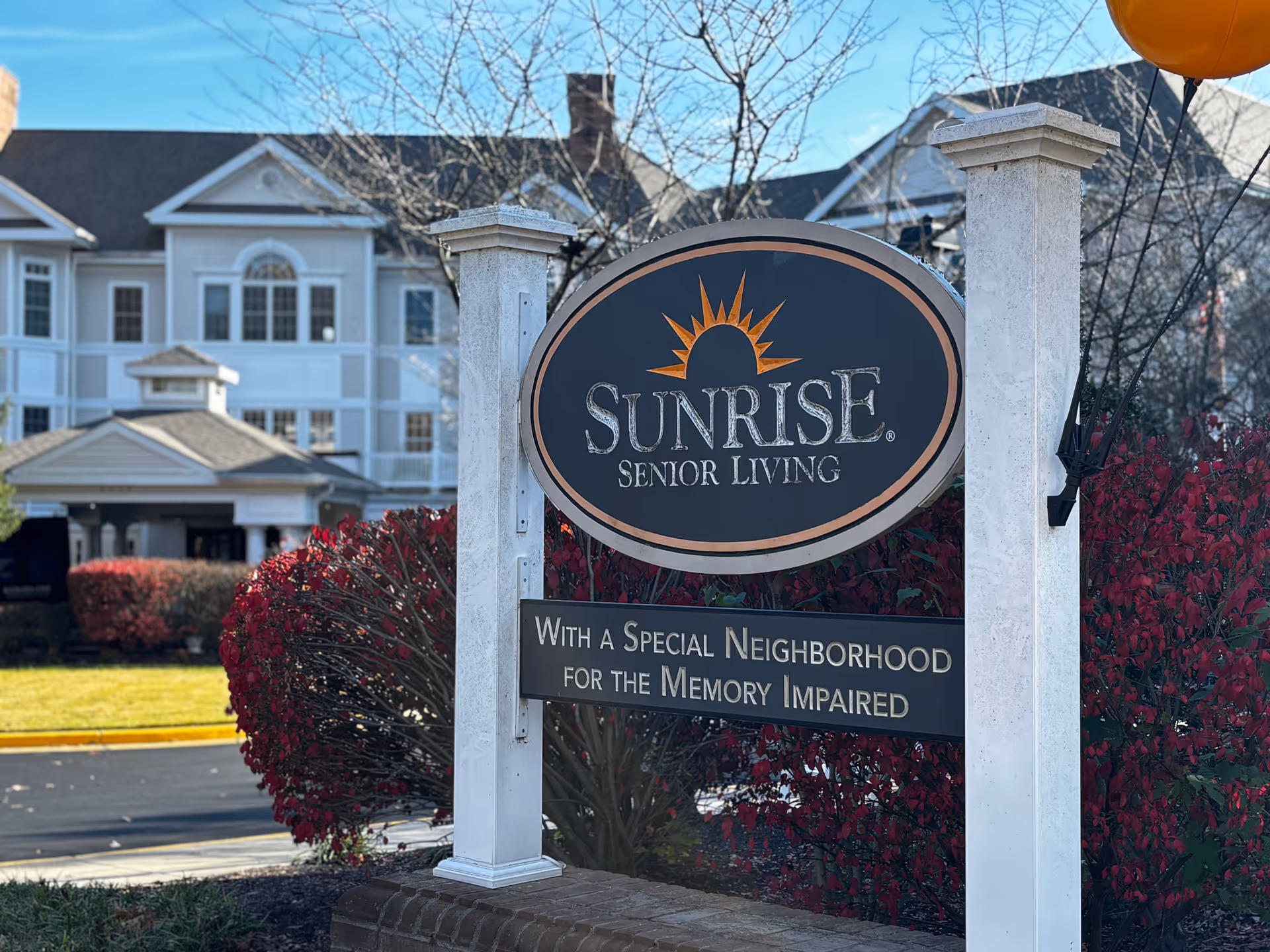 Outdoor view of a Sunrise Senior Living sign with a message indicating a special neighborhood for the memory impaired, set against a backdrop of a large building and red bushes under a clear blue sky.