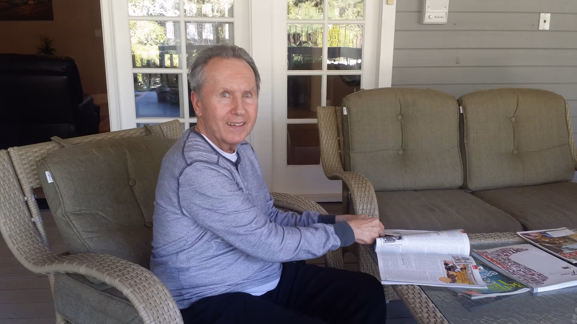 An elderly man sitting on a cushioned wicker chair on a covered patio, reading a magazine placed on a glass-top table next to a cushioned wicker sofa. Behind him are glass doors leading inside the building.