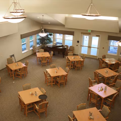 A spacious dining room with multiple square wooden tables, each surrounded by four chairs. The room has large windows on one side, letting in natural light, and a grand piano in the corner near the windows. There are modern hanging light fixtures from the ceiling and double glass doors leading outside.