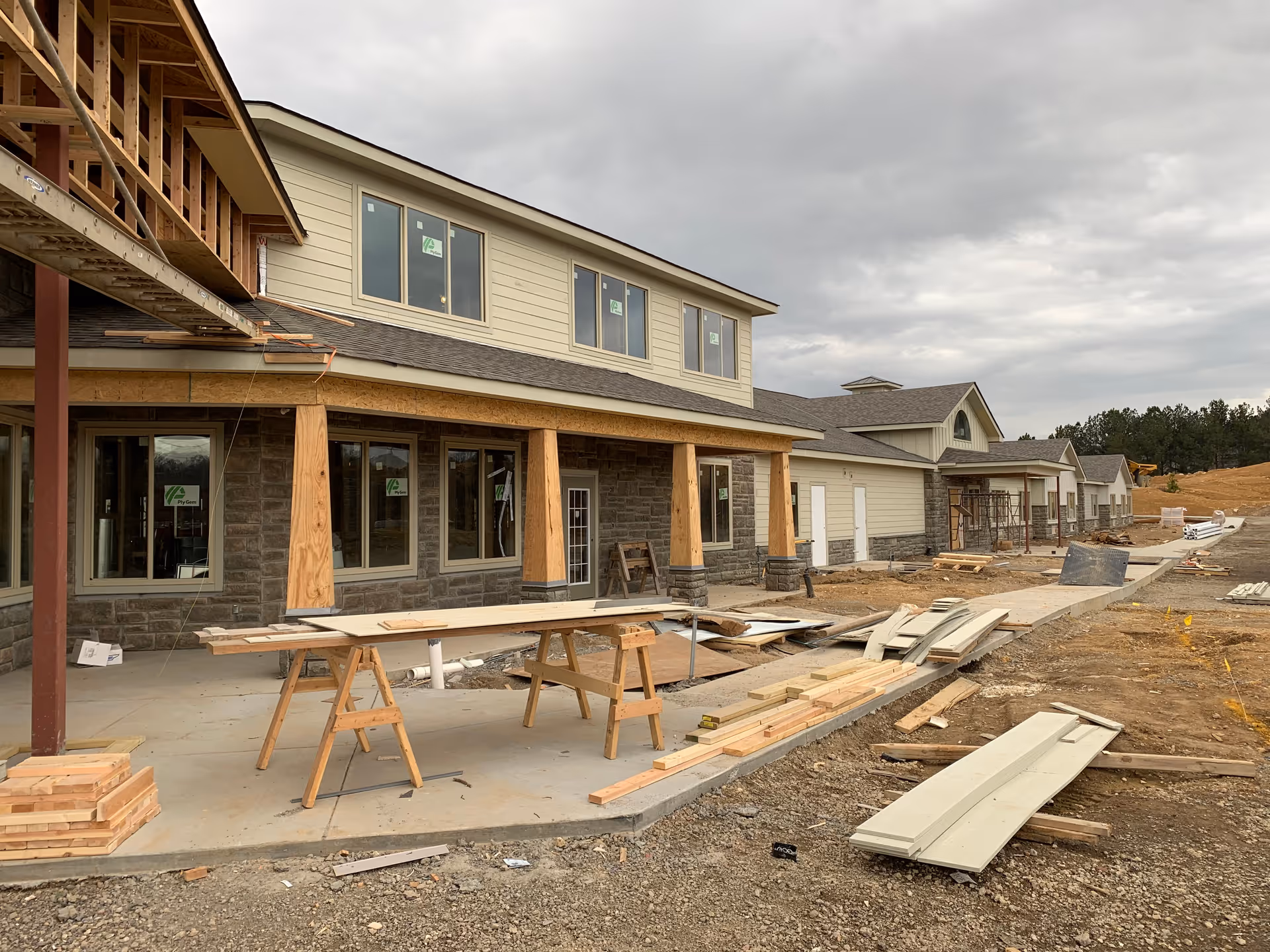 A senior living facility under construction with a two-story building featuring large windows and wooden support beams. Construction materials and tools are scattered around the site, with a cloudy sky overhead.