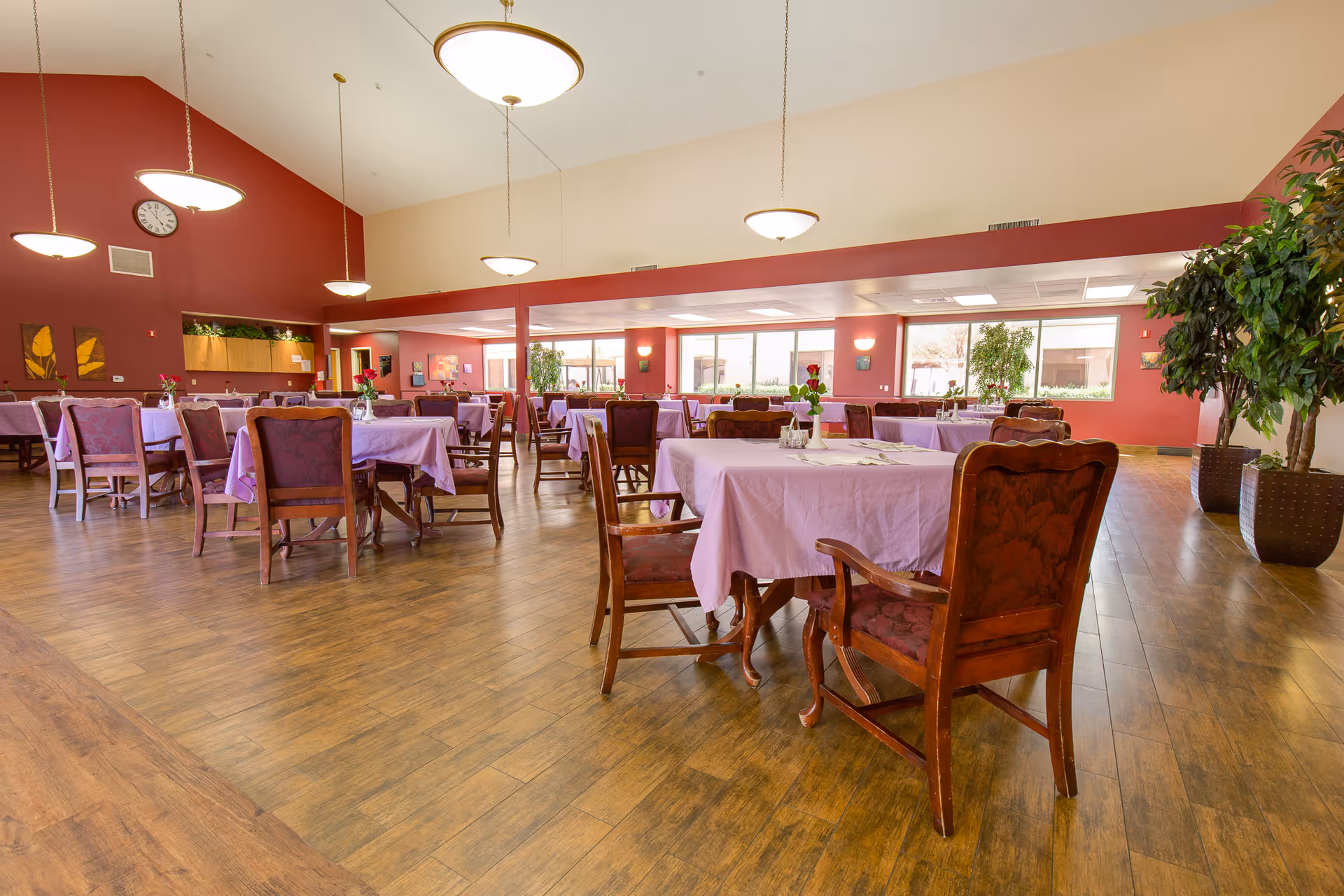 A spacious dining room with multiple tables covered in light purple tablecloths, each surrounded by wooden chairs with cushioned seats. The room has large windows allowing natural light, wooden flooring, red accent walls, hanging ceiling lights, and several potted plants.