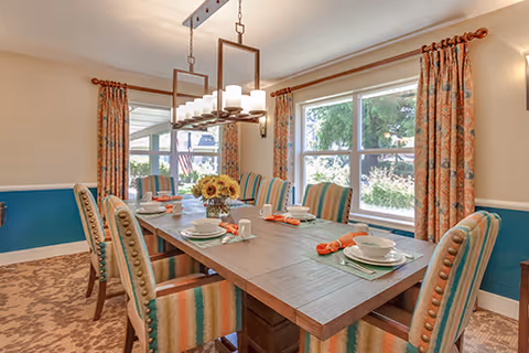 A bright dining room with a wooden table set for six, featuring striped upholstered chairs, a centerpiece of sunflowers, large windows with patterned curtains, and a modern rectangular chandelier overhead.