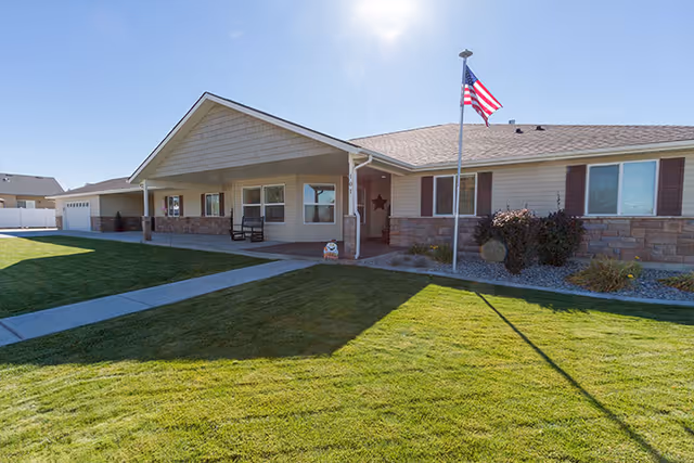 Single-story residential building with beige siding and stone accents, a covered front porch with seating, an American flag on a flagpole in the front yard, and a well-maintained green lawn under a clear blue sky.