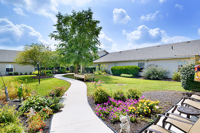 A sunny outdoor garden area with a curved concrete walkway surrounded by colorful flower beds, green shrubs, and trees. There are benches along the path and single-story buildings with light-colored siding in the background under a partly cloudy blue sky.