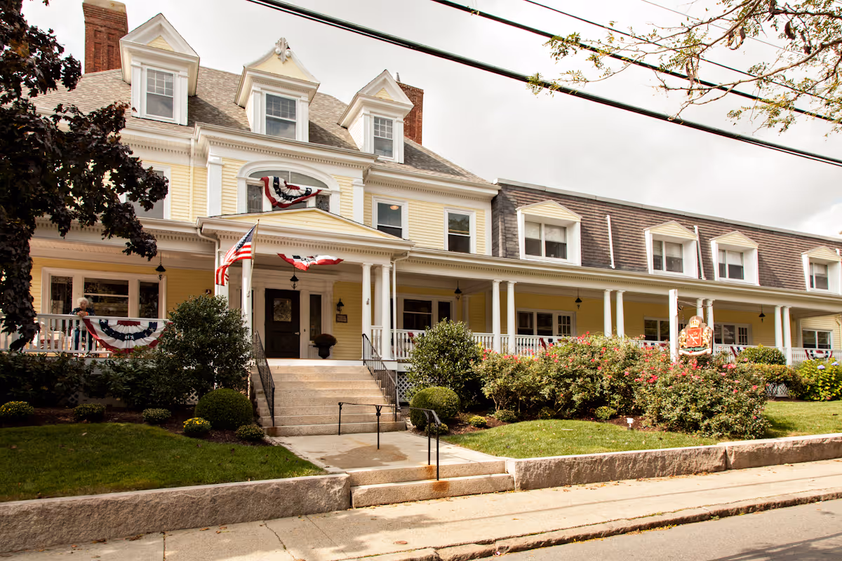 Yellow Victorian-style nursing center with a wide covered front porch, steps from the sidewalk, American flags, and landscaped shrubs.