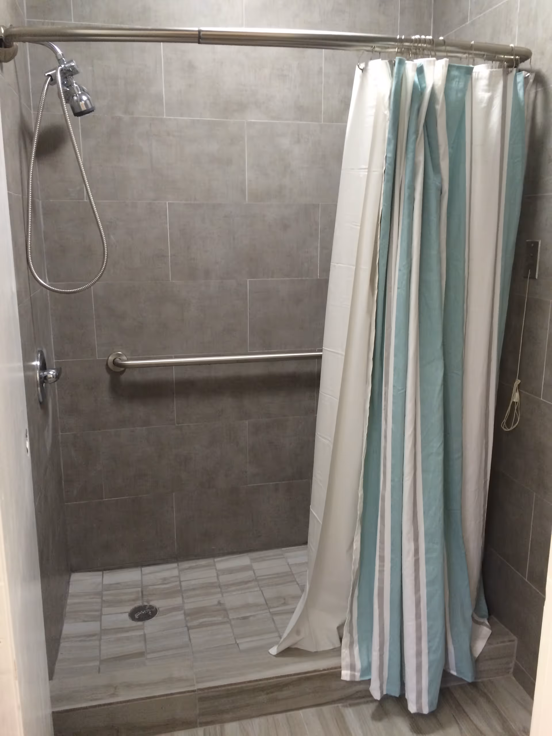 A tiled walk-in shower with gray wall tiles and a light gray floor. The shower has a silver handheld showerhead attached to a hose, a horizontal grab bar on the back wall, and a curved shower curtain rod with a white and light blue striped shower curtain partially closed.