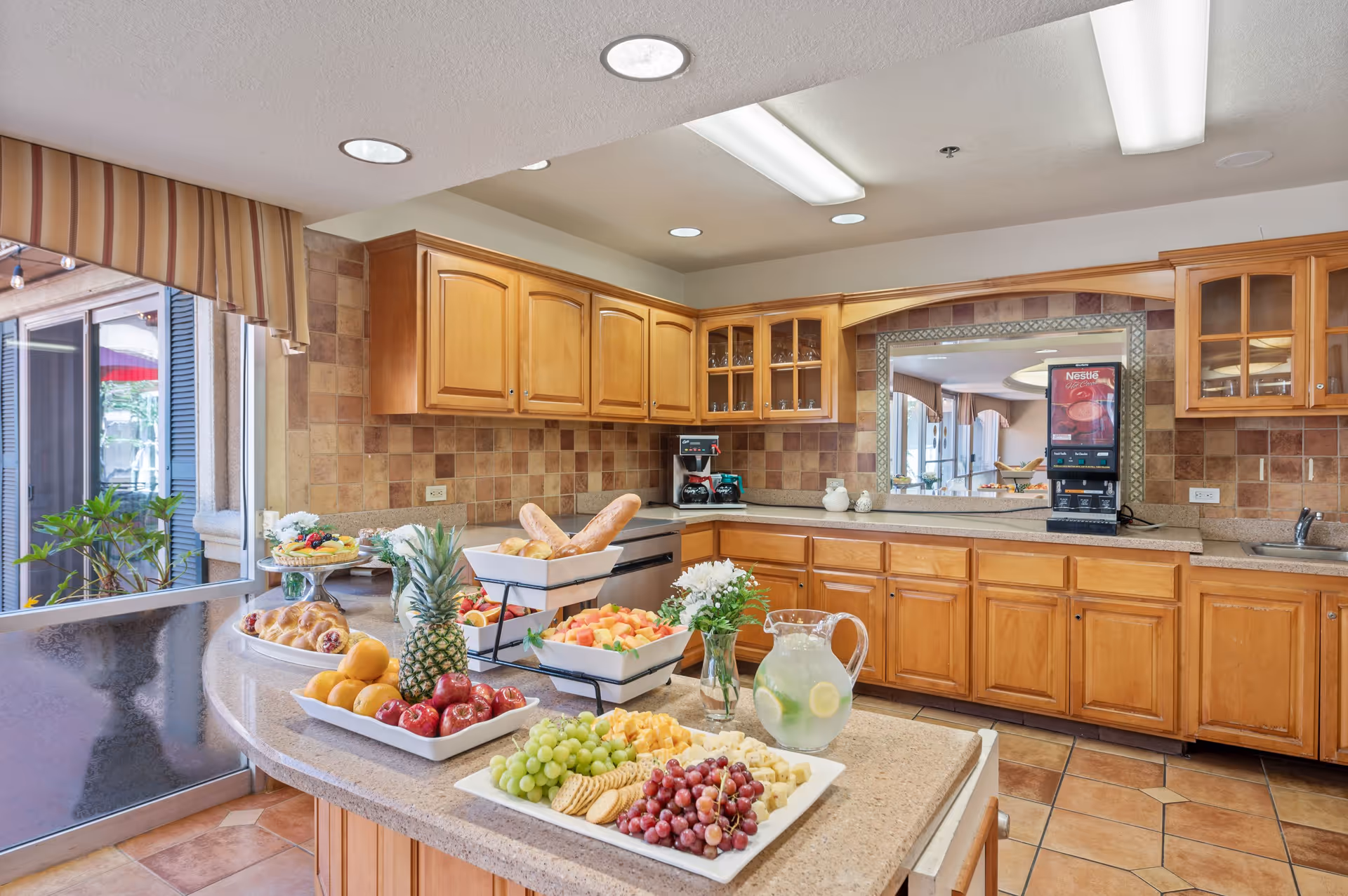 A bright kitchen area with wooden cabinets and tiled backsplash. The countertop is filled with various fresh fruits, bread, pastries, and a pitcher of lemonade with lemon slices. There is a coffee machine and a Nestle beverage dispenser on the counter. A large window with striped valance overlooks an outdoor area with plants.