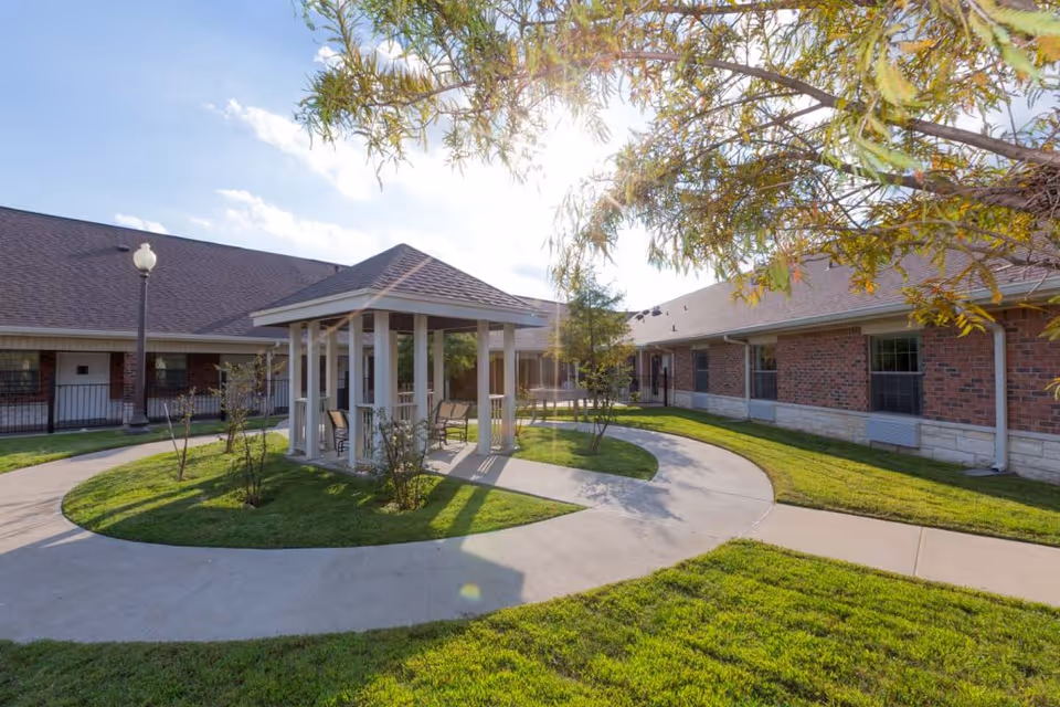 Outdoor courtyard area at Legend Oaks Healthcare and Rehabilitation - Ennis featuring a small gazebo with chairs inside, surrounded by a circular concrete walkway, green grass, and trees with sunlight filtering through.