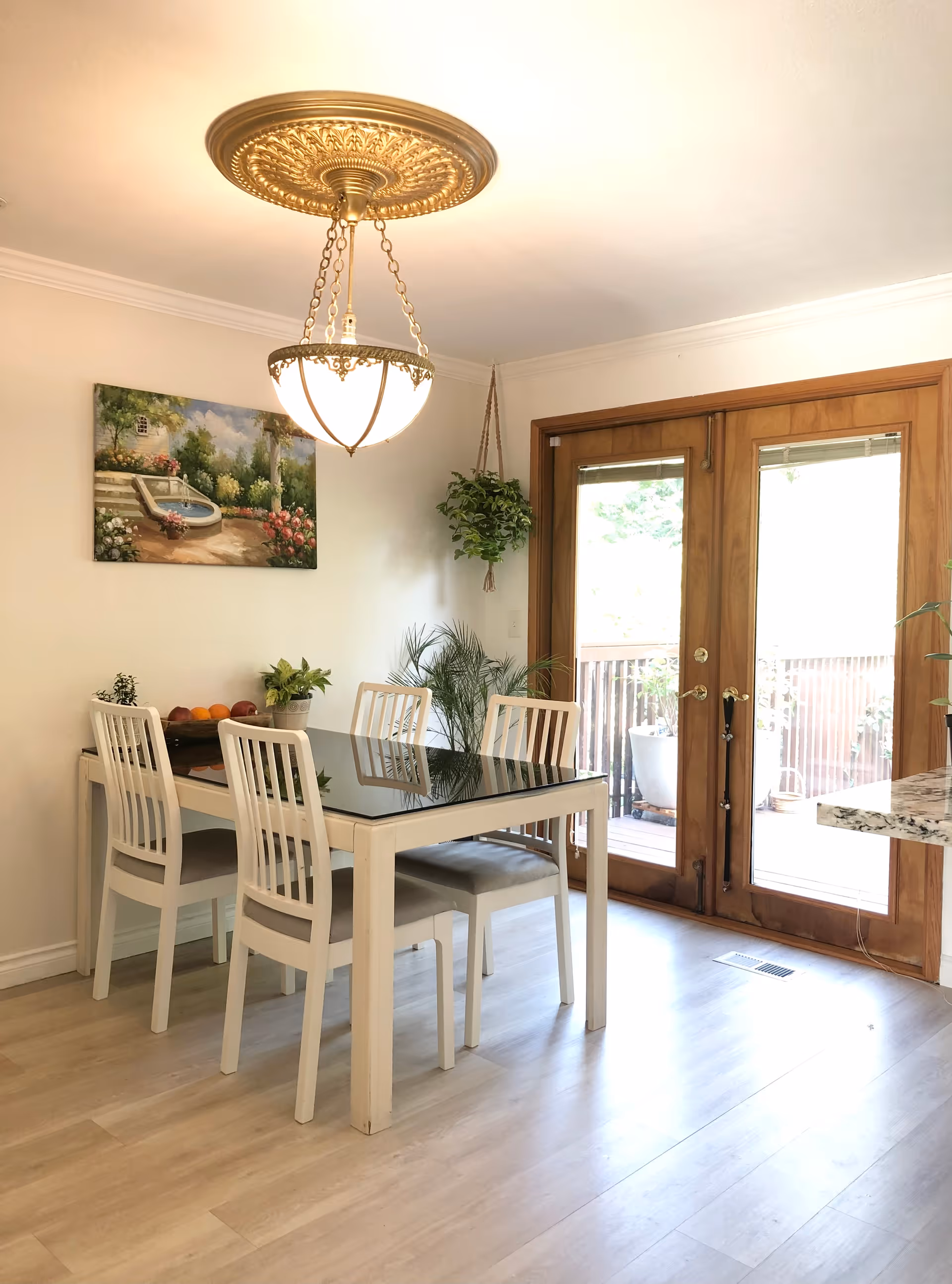 A bright dining area with a white table and four white chairs with gray cushions. A decorative gold chandelier hangs above the table. There is a painting of a garden scene on the wall and several potted plants, including a hanging plant near wooden double doors that lead to an outdoor deck.
