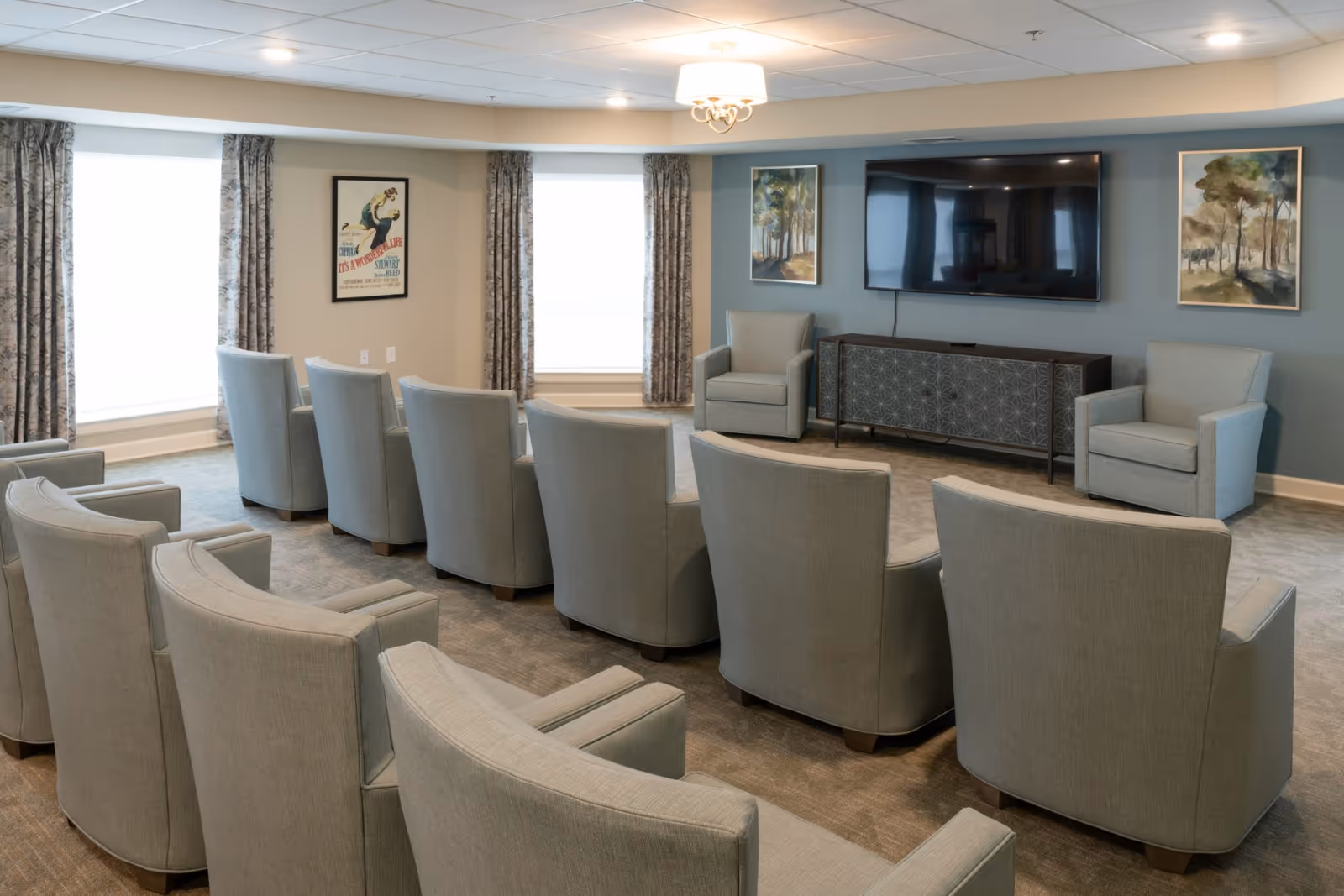A spacious living room with multiple light gray upholstered armchairs arranged in rows facing a large flat-screen TV mounted on a blue accent wall. Below the TV is a decorative console table. The room has two large windows with patterned curtains, framed artwork on the walls, and a ceiling light fixture.