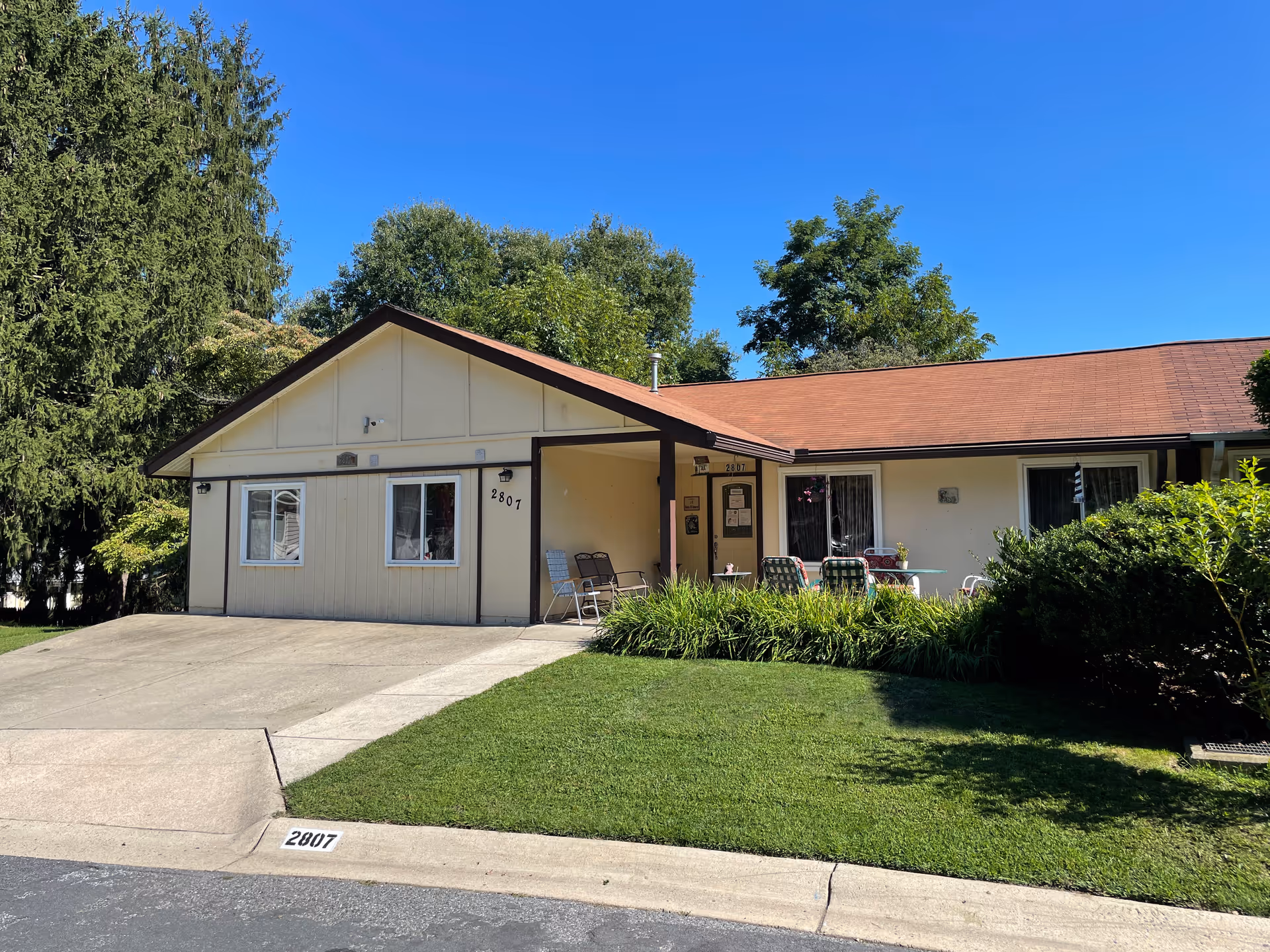 Single-story assisted living house with a driveway, front lawn, and patio chairs under a clear blue sky.