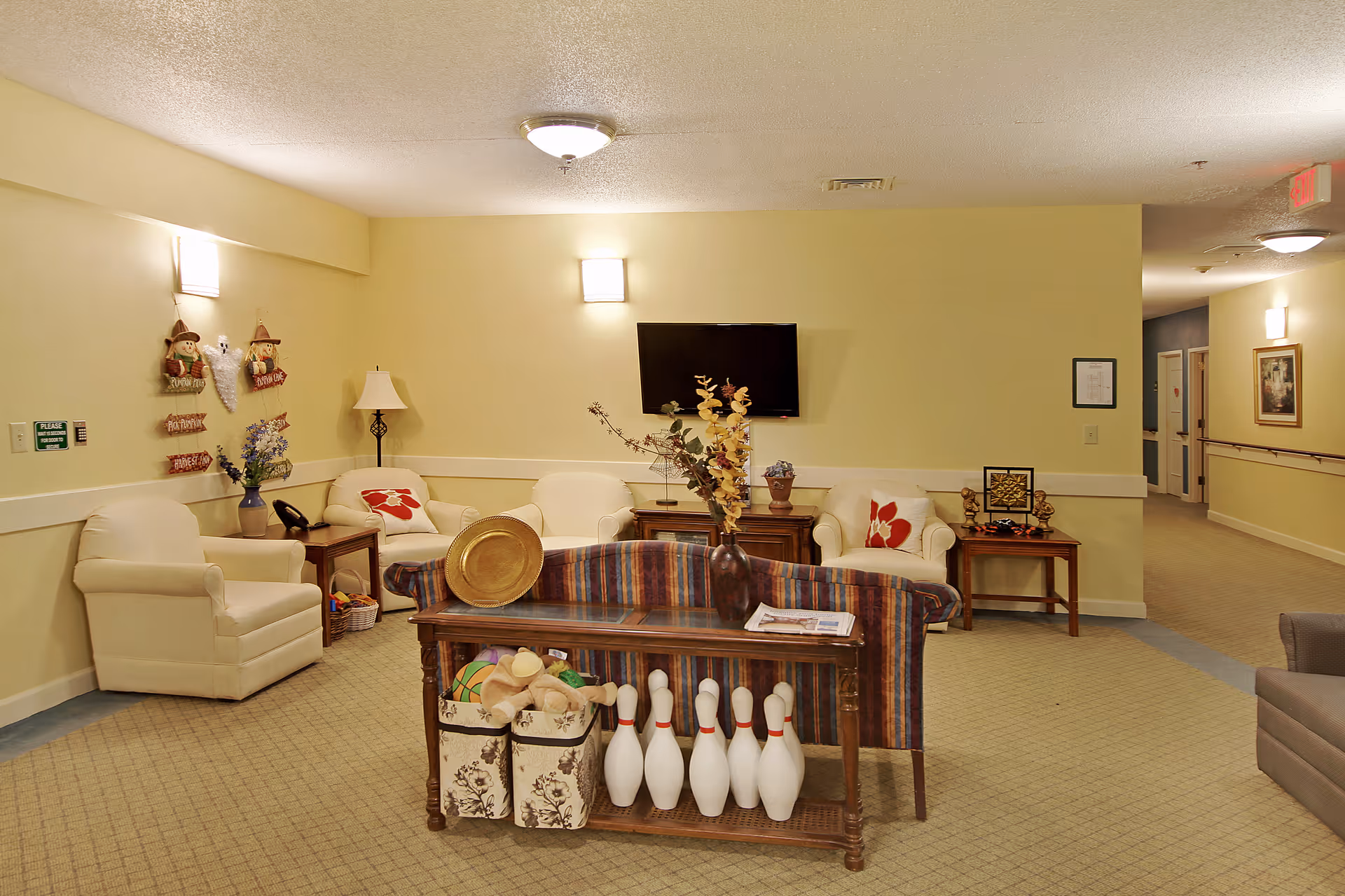 Cozy senior living common room with armchairs, a wall-mounted TV, a striped sofa, and a console table holding decorative items and bowling pins.