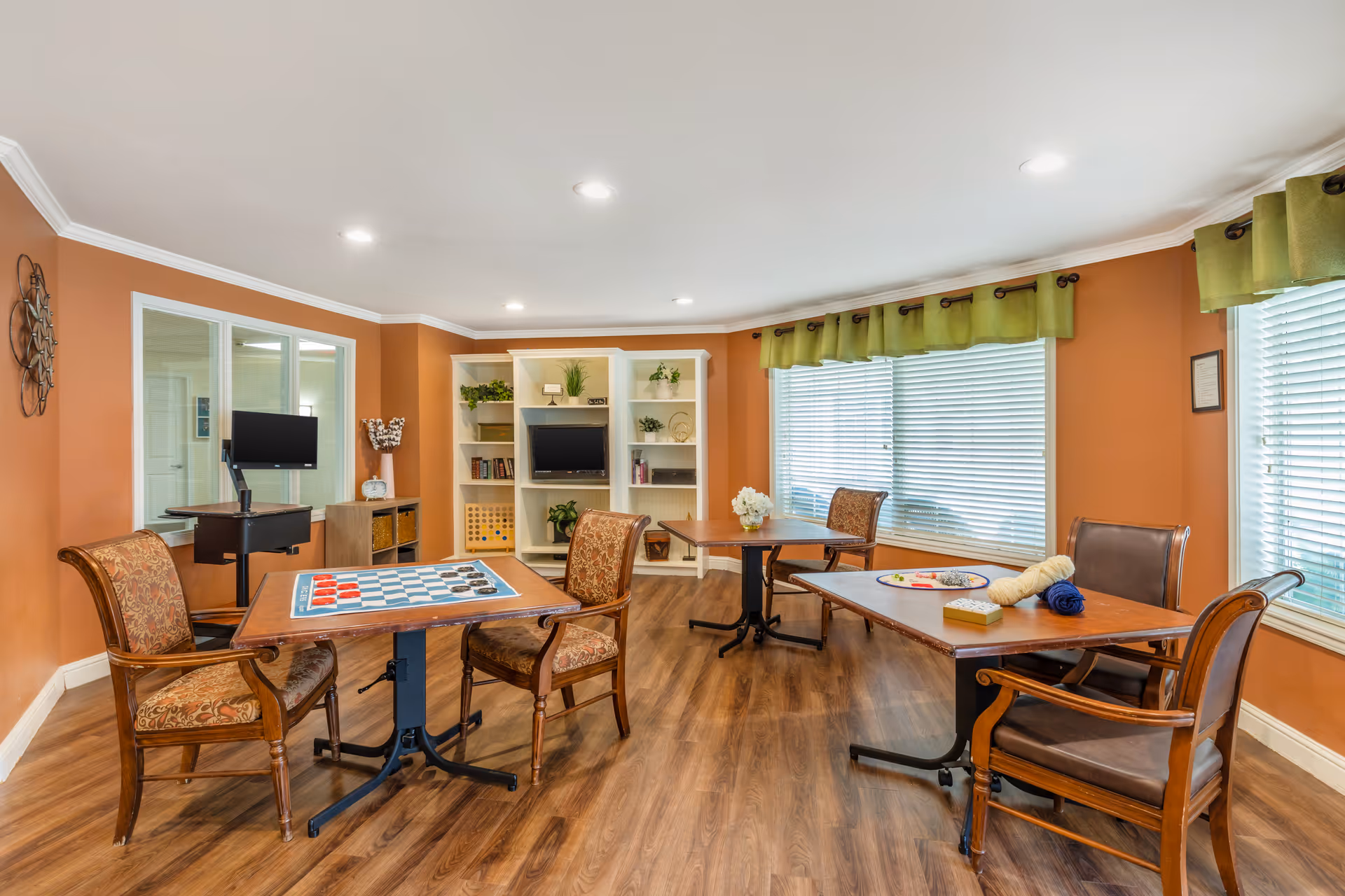 Bright senior living activity room with tables and chairs, a TV and shelving against orange walls and large windows.