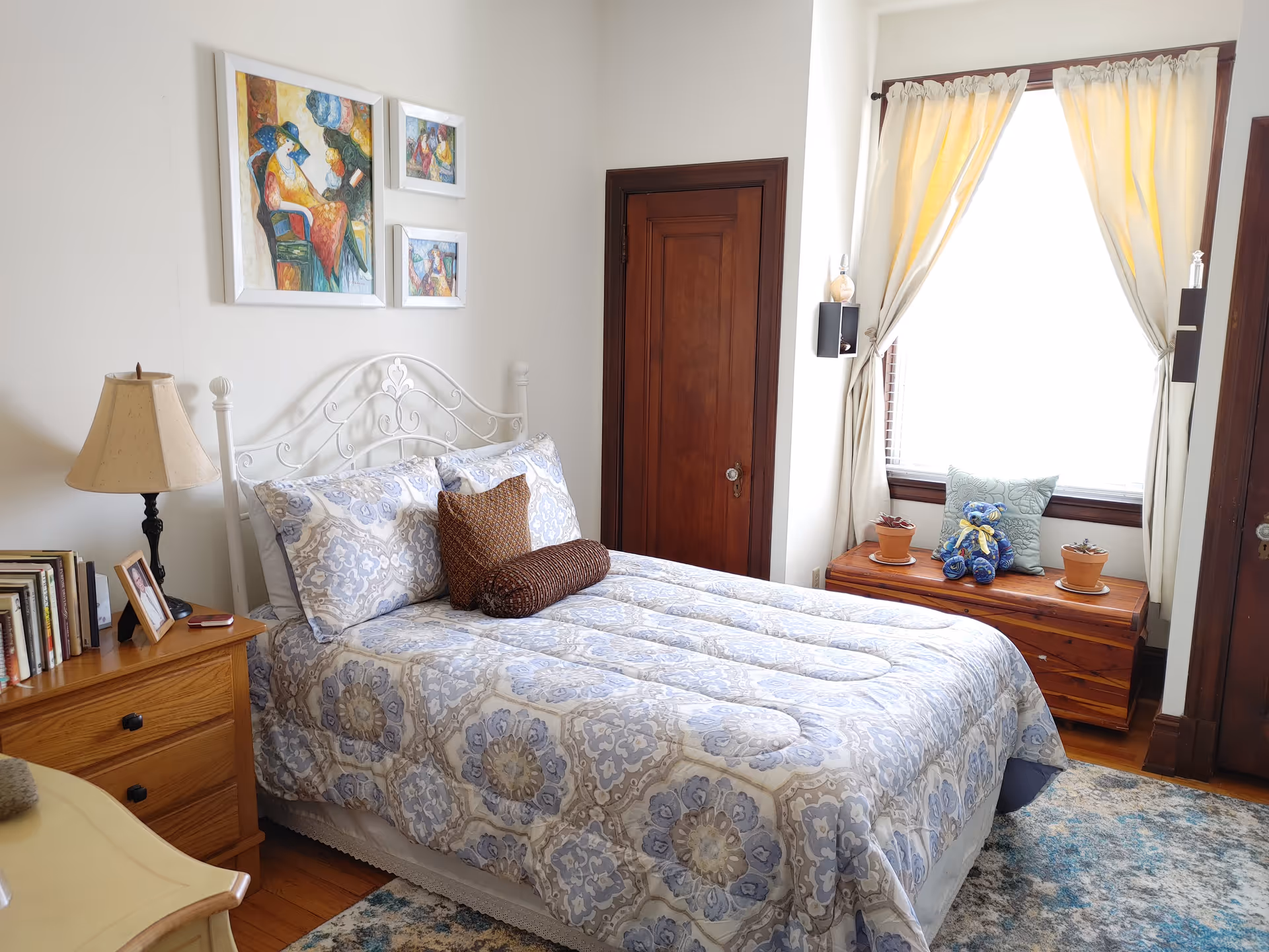 A cozy bedroom with a neatly made bed featuring a patterned blue and beige comforter and pillows. There is a wooden nightstand with a lamp, books, and a framed photo beside the bed. On the wall above the bed are three framed colorful paintings. A wooden chest sits under a window with light curtains, decorated with a blue teddy bear, a pillow, and two small potted plants. The room has wooden doors and a patterned rug on the floor.