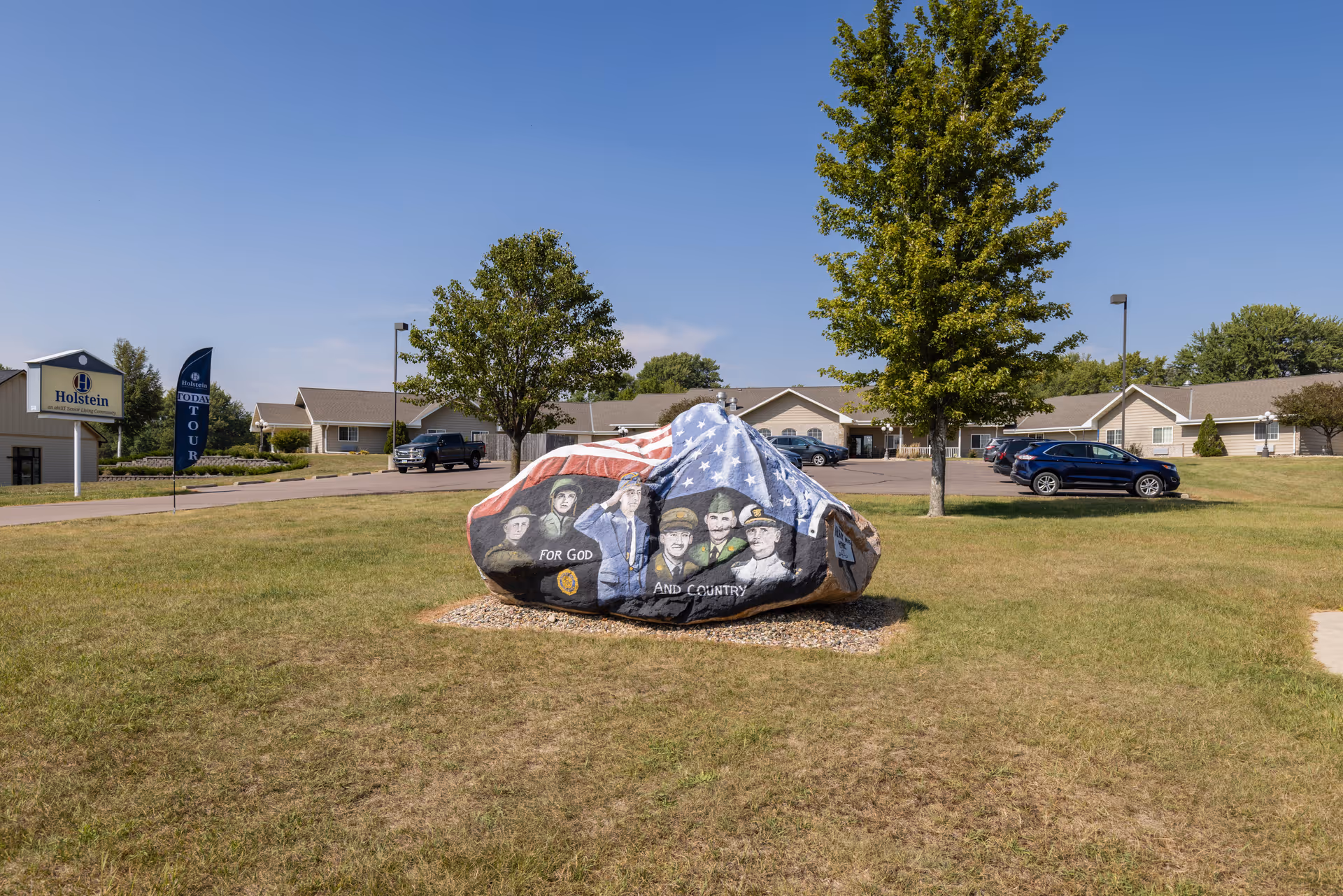 Outdoor view of Holstein Senior Living facility with a large painted rock in the foreground featuring an American flag and portraits of military personnel with the words 'For God and Country'. The building and parking area with cars are visible in the background under a clear blue sky.