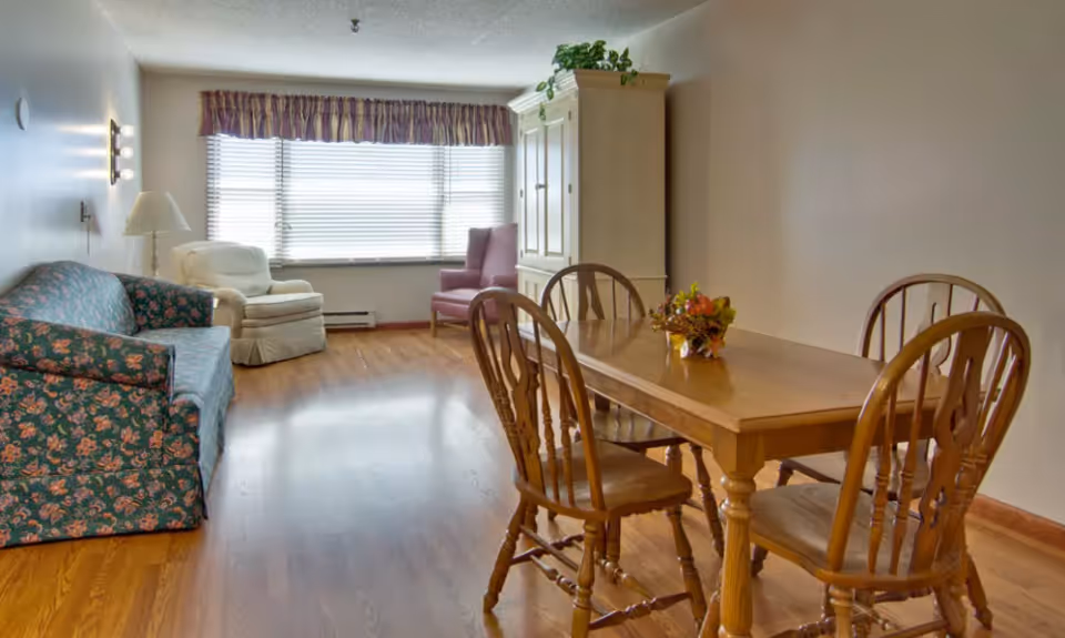 A well-lit living and dining area with a wooden dining table and four chairs in the foreground. In the background, there is a floral-patterned sofa, a white armchair, a purple armchair, and a large window with blinds and a valance. A white cabinet with a plant on top is against the wall.