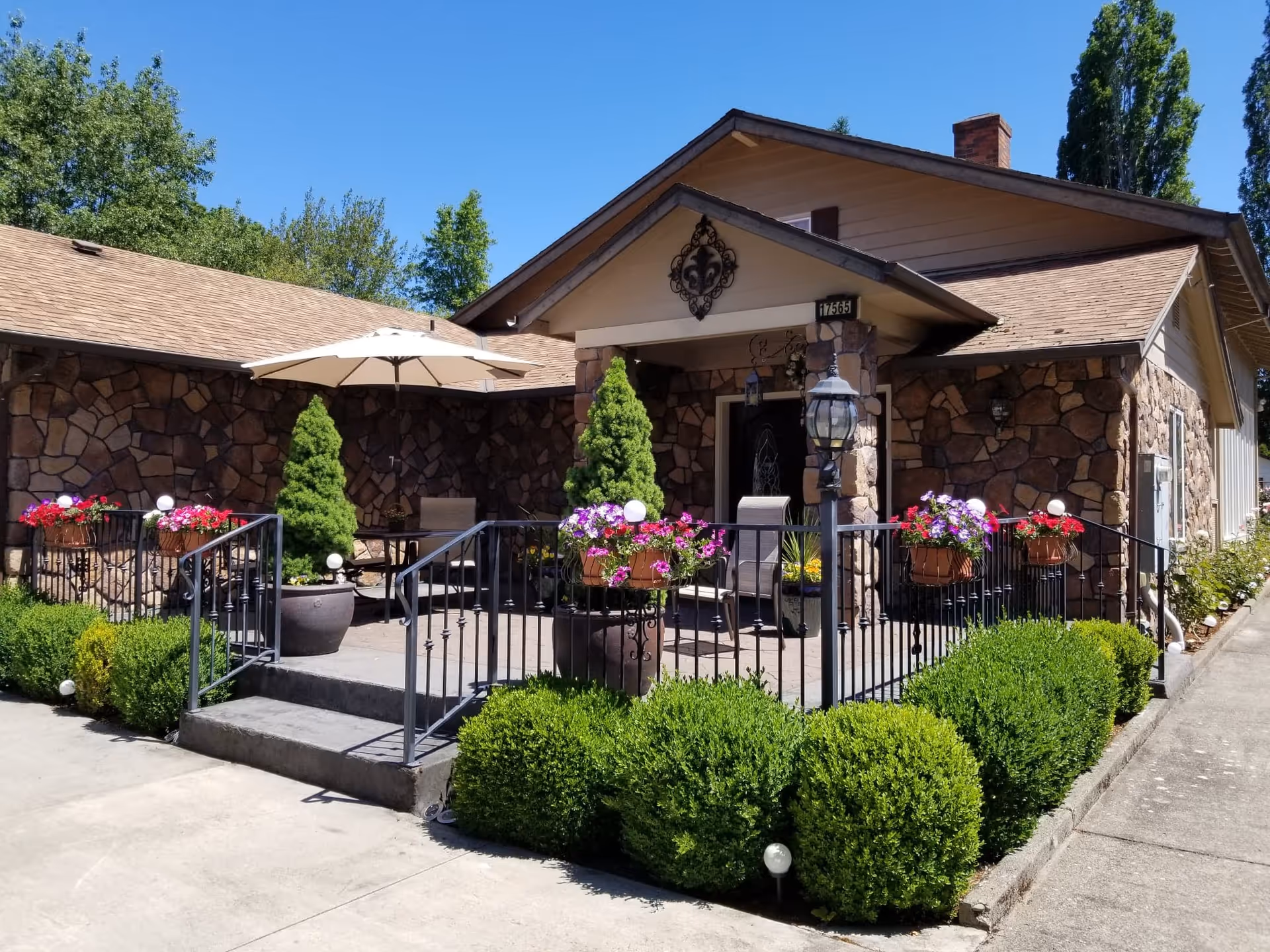 Front exterior view of a single-story senior living facility with stone facade, a small porch with outdoor seating, potted plants, flowers, and a large umbrella. The area is surrounded by neatly trimmed bushes and trees under a clear blue sky.